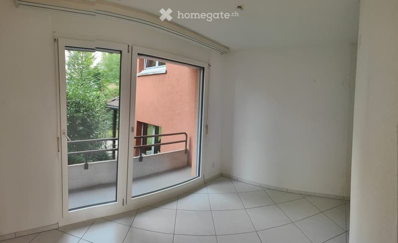 Empty room with tiled floor, white walls, and large sliding glass doors leading to a balcony with metal railing and greenery visible outside.