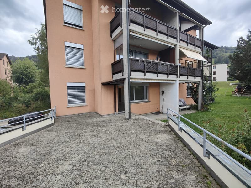 Two-story residential building with balconies, beige facade, and multiple windows. A car is parked near the entrance, and there is a grassy area with outdoor furniture visible.