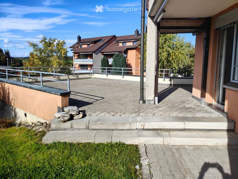 Balcony with concrete floor, metal railing, steps leading down to a paved area, and a small garden patch with grass and rocks.
