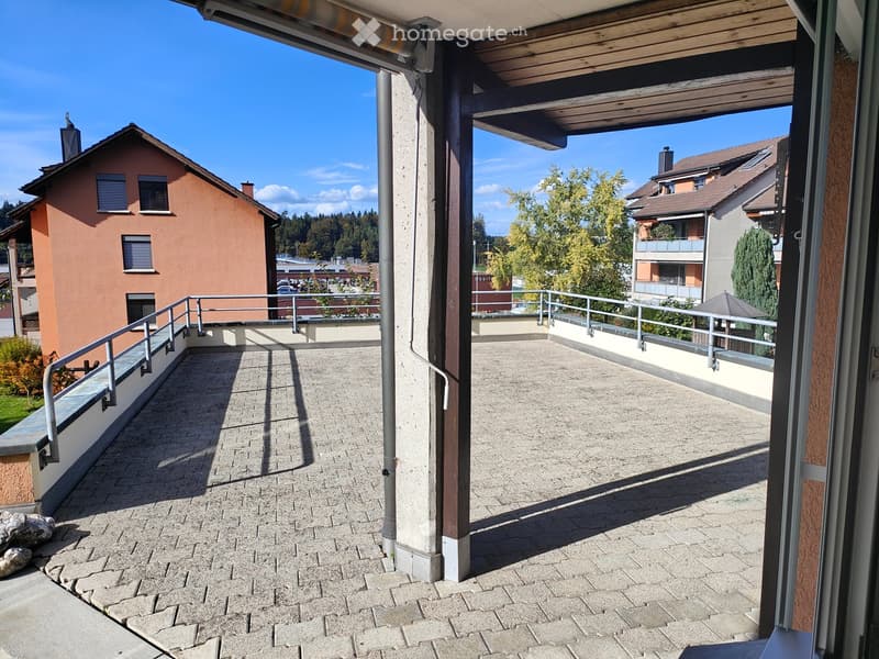 Balcony with brick pavement, metal railing, and wooden roof, overlooking neighboring houses and trees.