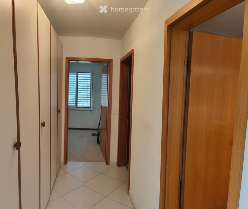A clean, well-lit hallway with tiled flooring, wooden doors, and a visible room at the end with windows and blinds.