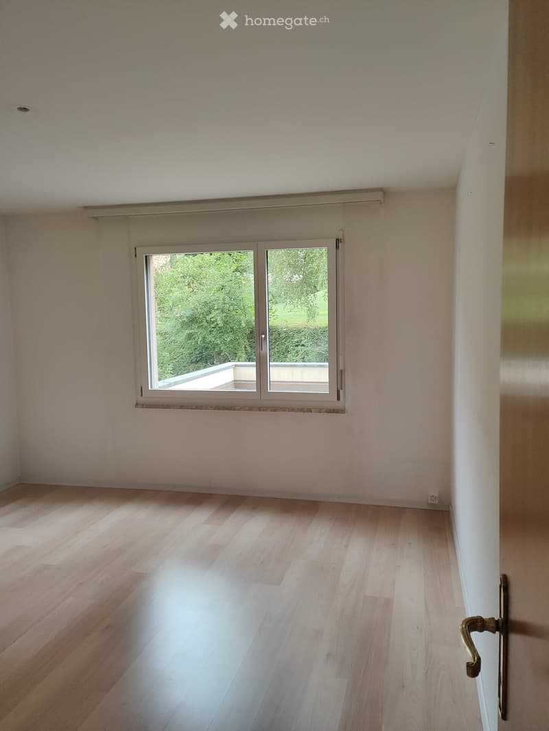 Empty room with light wooden flooring, white walls, and a large window with a view of greenery outside.
