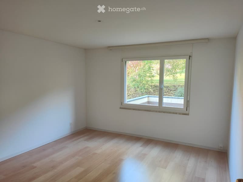 Empty room with light wooden flooring and white walls, featuring a large window with a view of a garden area.