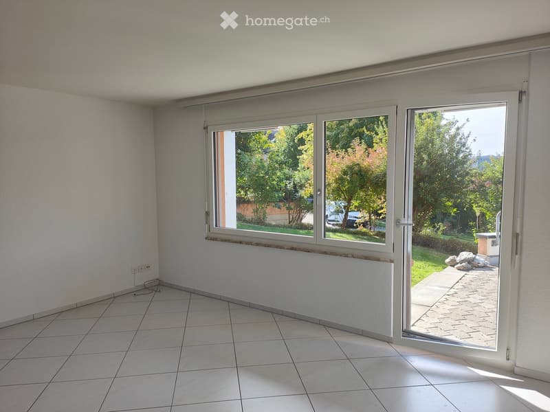 Empty room with tiled floor, white walls, large sliding glass doors leading to a garden area with trees and a paved pathway.