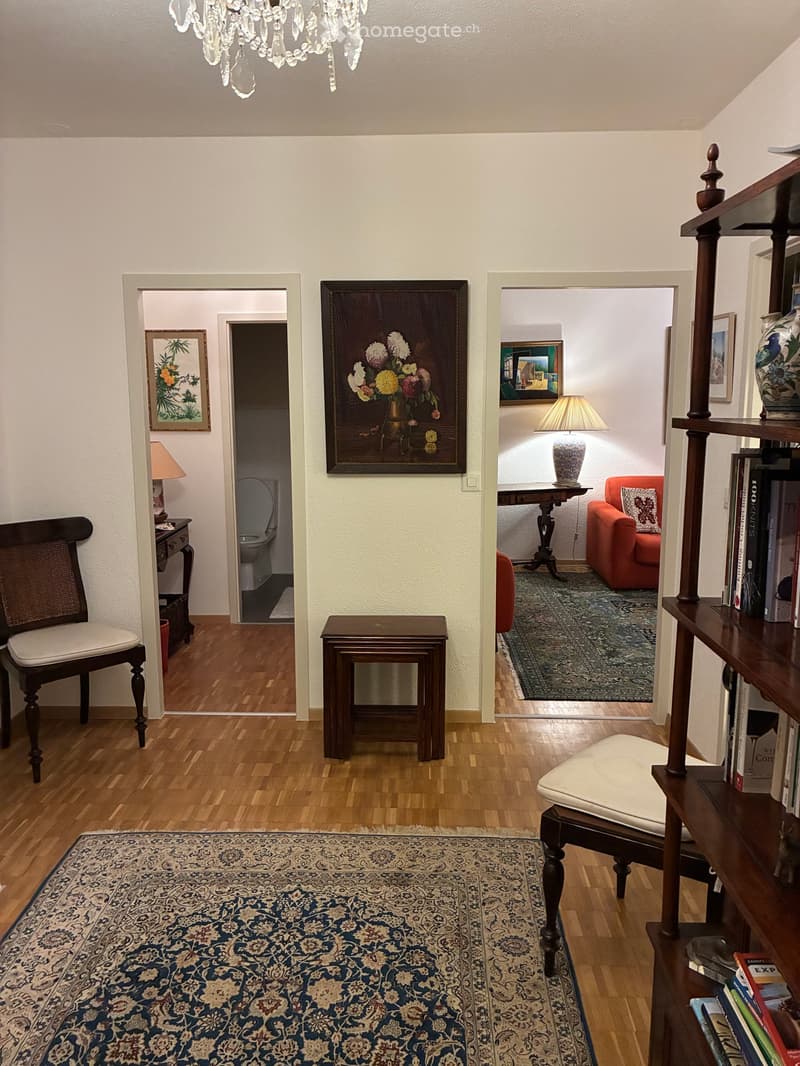 Elegant hallway with wooden flooring, chandelier, and multiple rooms visible, including a living area with a red armchair, a small table, and a bookshelf, as well as a bathroom with a toilet and a small table with a lamp.