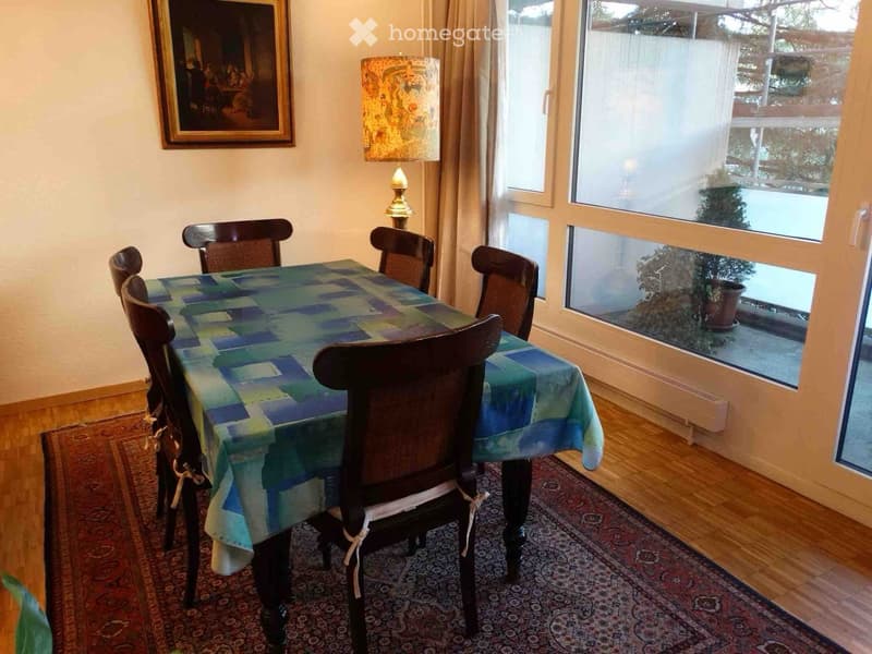 Dining room with a wooden table covered in a blue and green tablecloth, surrounded by wooden chairs, a lamp on a side table, and a painting on the wall. Large windows offer a view of the garden.