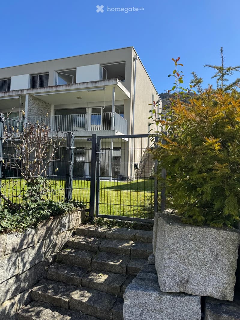 Modern two-story house with a raised ground floor, large windows, and a balcony. The house is surrounded by a well-maintained garden with stone steps leading up to the entrance.