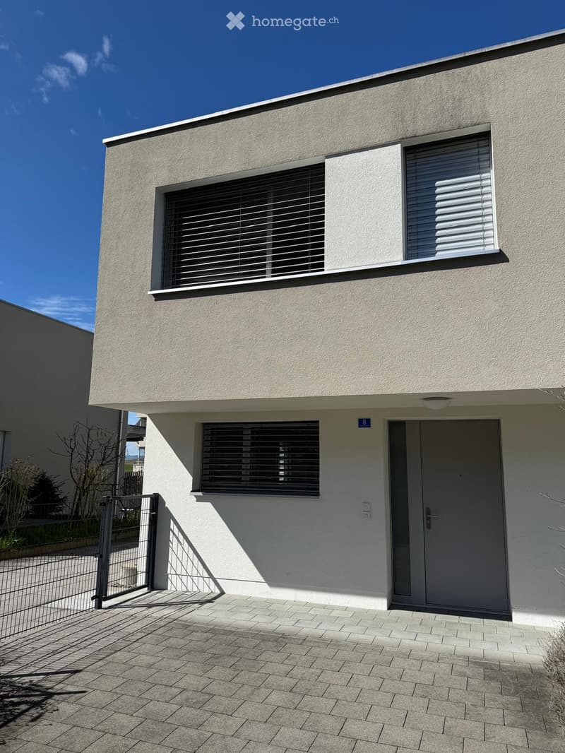 Modern two-story house with light gray facade, large windows with blinds, and a gated entrance.