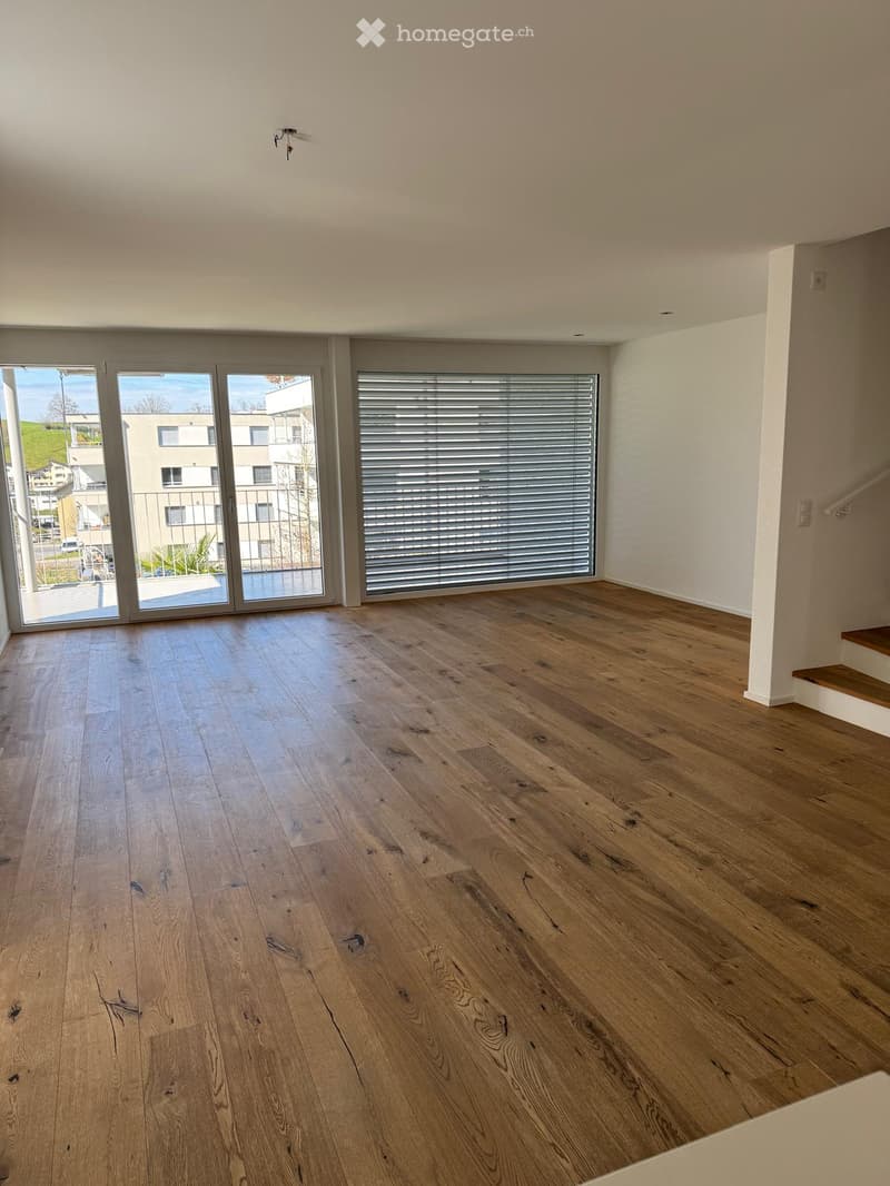 Empty room with light wooden flooring, large windows with blinds, and a staircase leading upwards.