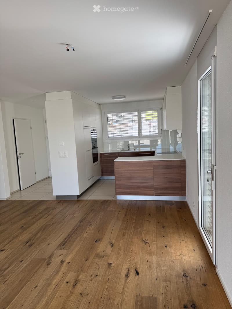 Empty kitchen with wooden flooring, white cabinets, and a central island. The space features large windows with blinds and a door leading to another room.