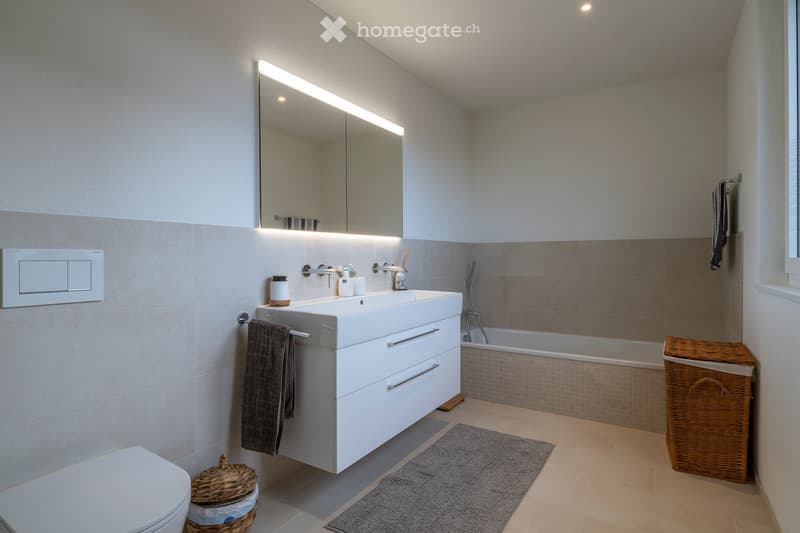 Modern bathroom with a double sink, large mirror with LED lighting, bathtub, and wicker baskets on the floor.