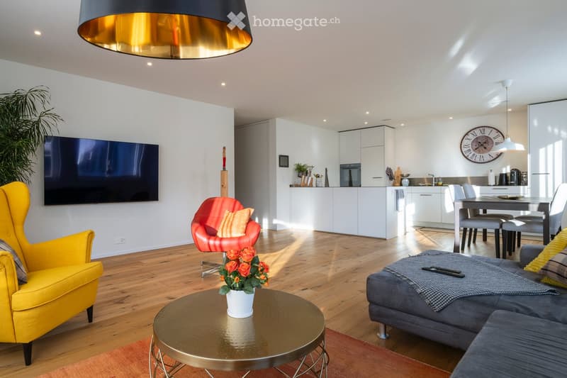 Modern living room with a grey sofa, yellow armchair, red chair, coffee table, dining table, and kitchen area with white cabinets and stainless steel appliances.