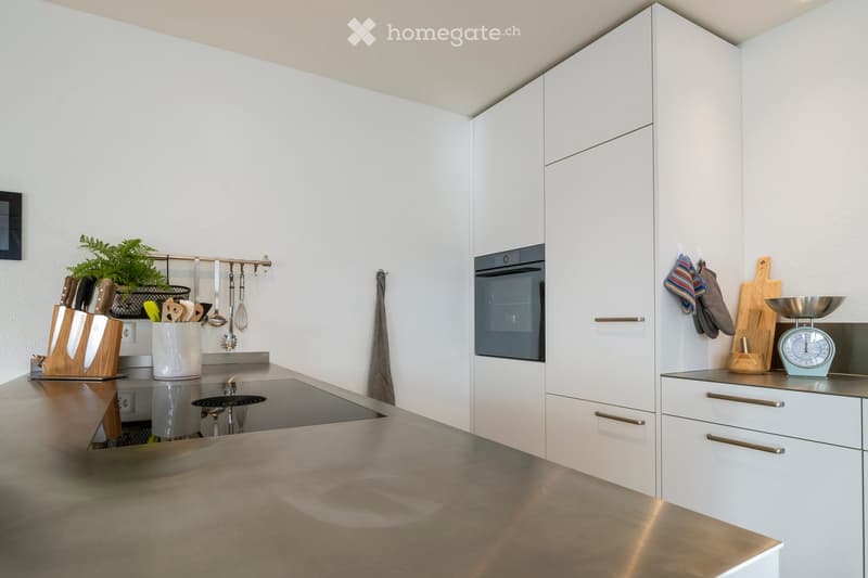 Modern kitchen with white cabinets, stainless steel countertop, built-in oven, knife block, hanging utensils, and a small plant on the counter.