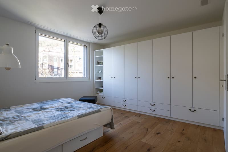 Modern bedroom with a large bed, white built-in wardrobe, a small black stool, and shelves with decorative items. The room features wooden flooring and large windows with a view of the street.
