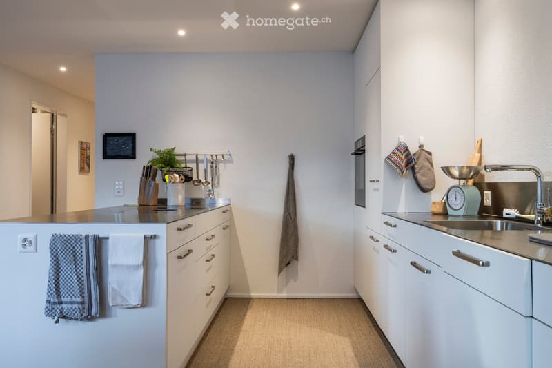 Modern kitchen with white cabinets, stainless steel appliances, and a central island. Features include a sink, oven, knife block, cutting board, and decorative towels.