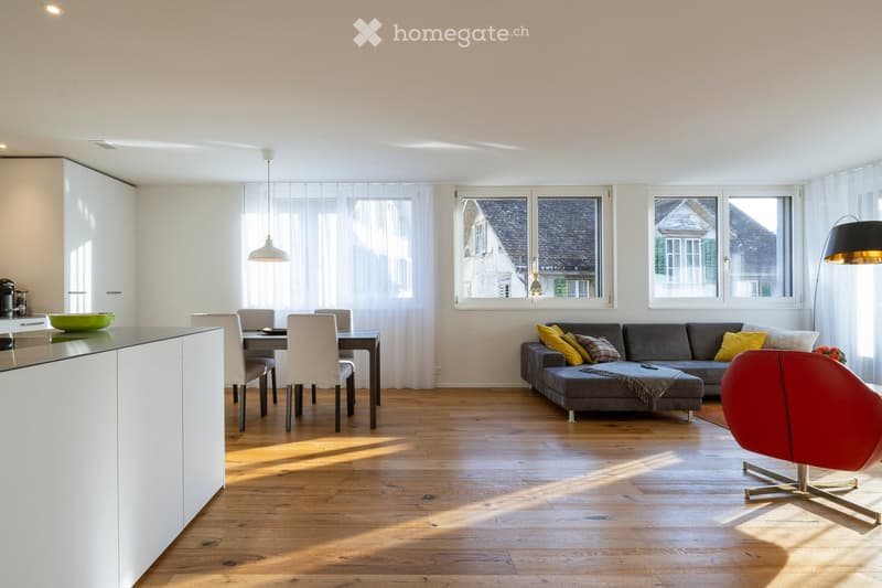 Open-plan living and dining area with wooden flooring, large windows, grey sectional sofa, red accent chair, dining table with chairs, and modern kitchen counter.