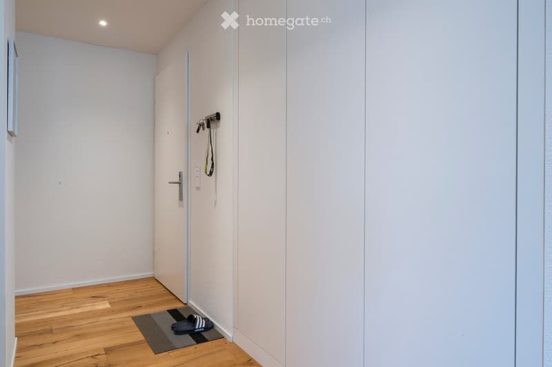 A narrow hallway with light-colored walls and wooden flooring, featuring a closed door, a small rug, and a pair of black sandals placed on the rug.