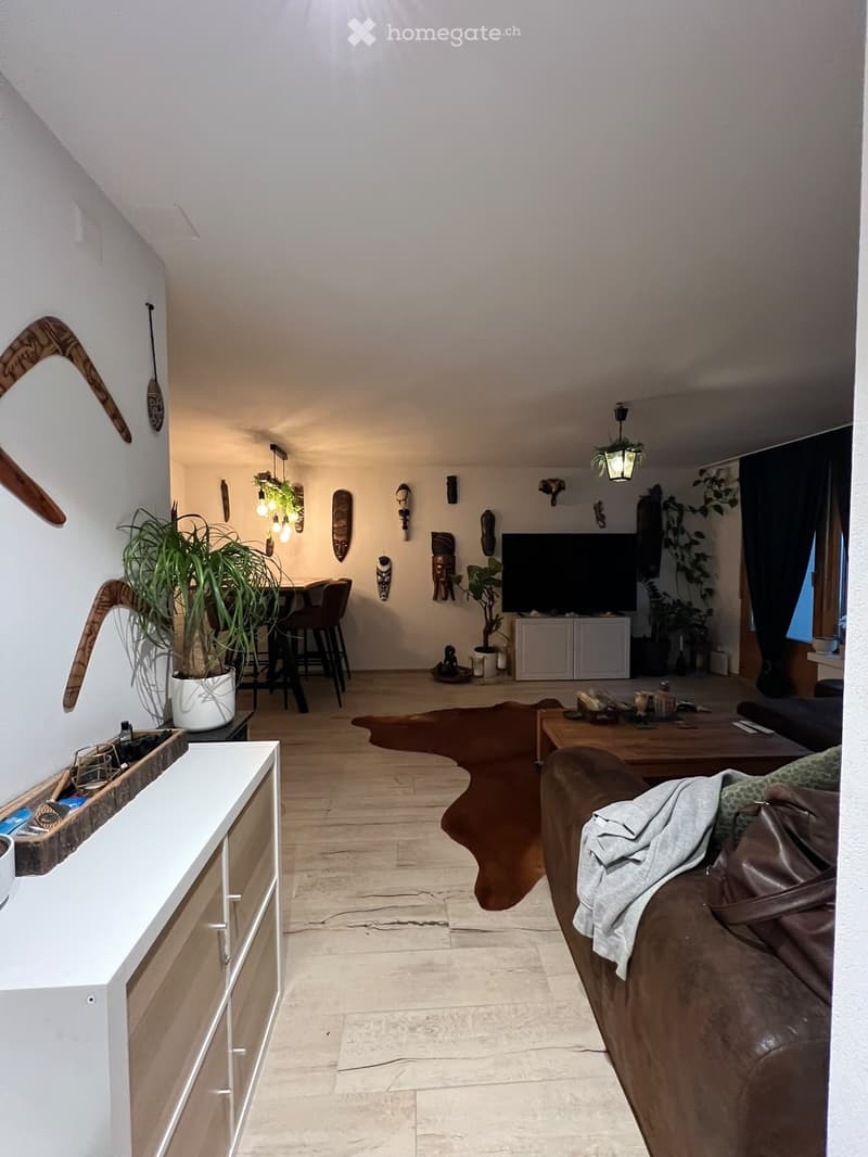 A modern living room with a brown leather sofa, coffee table, television, dining area with chairs and table, and decorative plants.