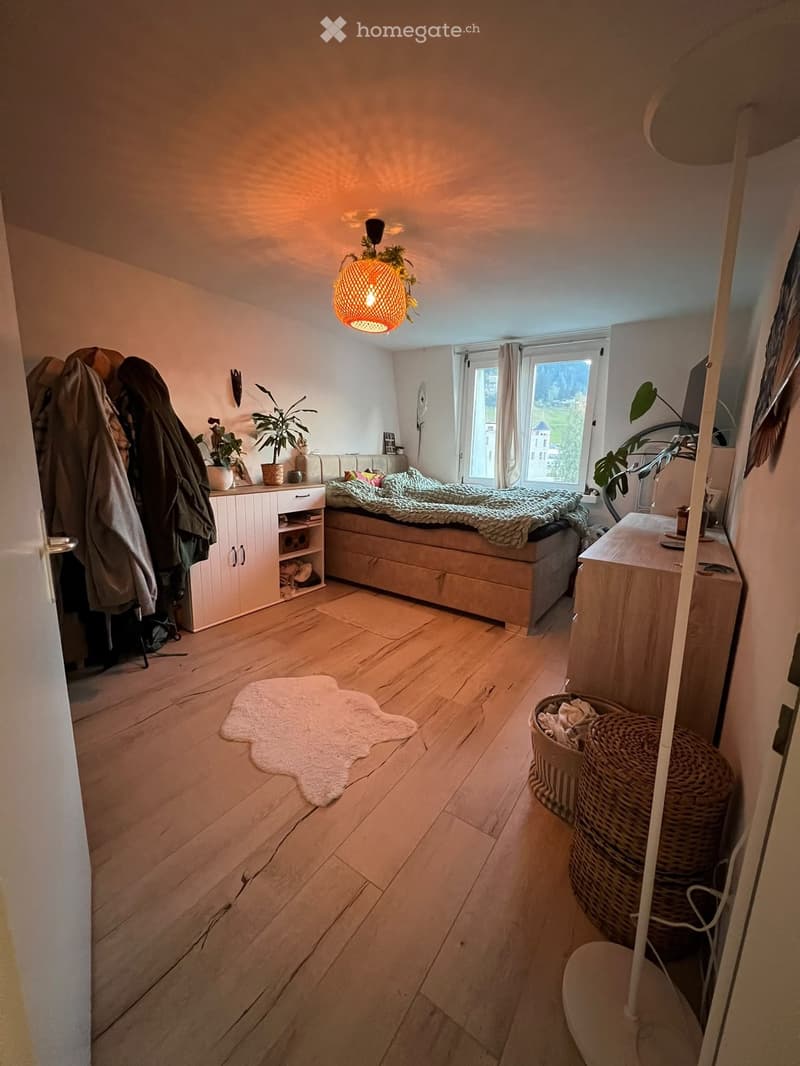Cozy bedroom with wooden flooring, a bed with a green blanket, a nightstand, a cabinet, a white rug, and decorative plants.