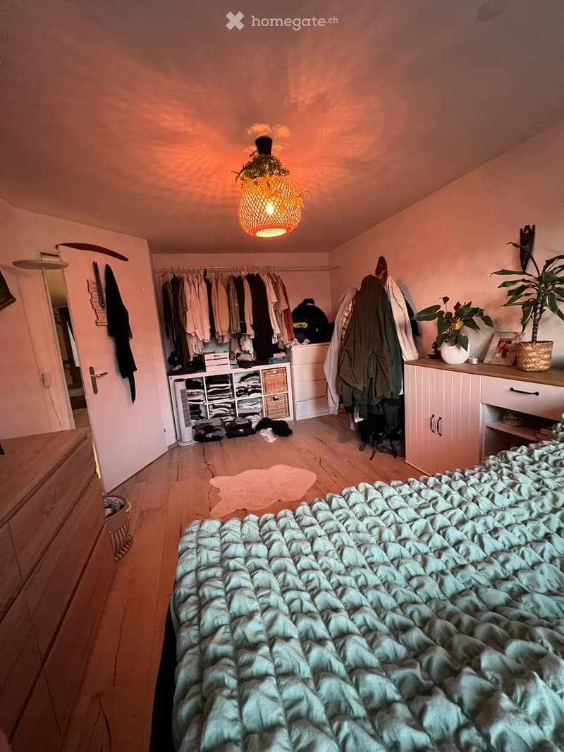 Cozy bedroom with a neatly made bed, wooden floor, wardrobe, dresser, and hanging clothes. Features a hanging woven lamp and potted plants.