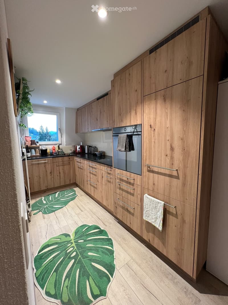 Modern kitchen with light wooden cabinets, stainless steel appliances, and a black countertop. Features include a built-in oven, refrigerator, coffee maker, toaster, and a green leaf-shaped rug on the floor.
