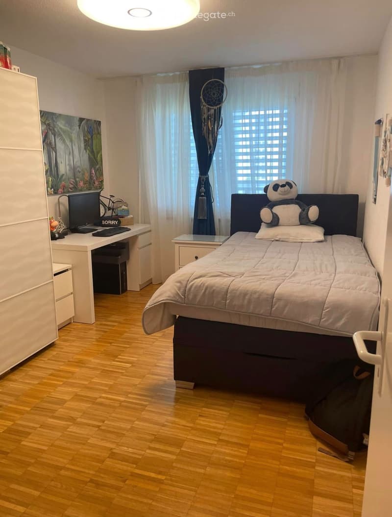 A modern bedroom with a double bed, desk, chair, and large window with curtains. The room features a wooden floor and a dreamcatcher hanging on the wall.