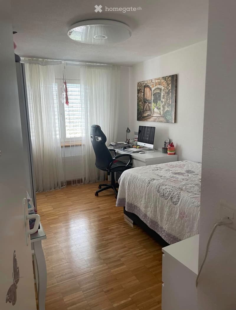 Cozy bedroom with wooden flooring, a double bed, a desk with a computer, a chair, and a large window with curtains. A painting hangs on the wall.