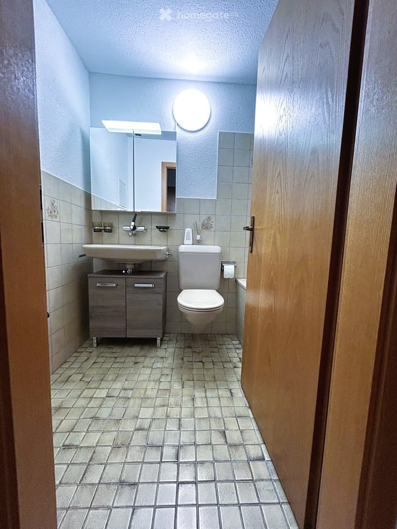 Bathroom with tiled floor and walls, a sink with a cabinet, a toilet, and a mirror above the sink. The room has a simple, clean design with a wooden door frame visible on the right side.