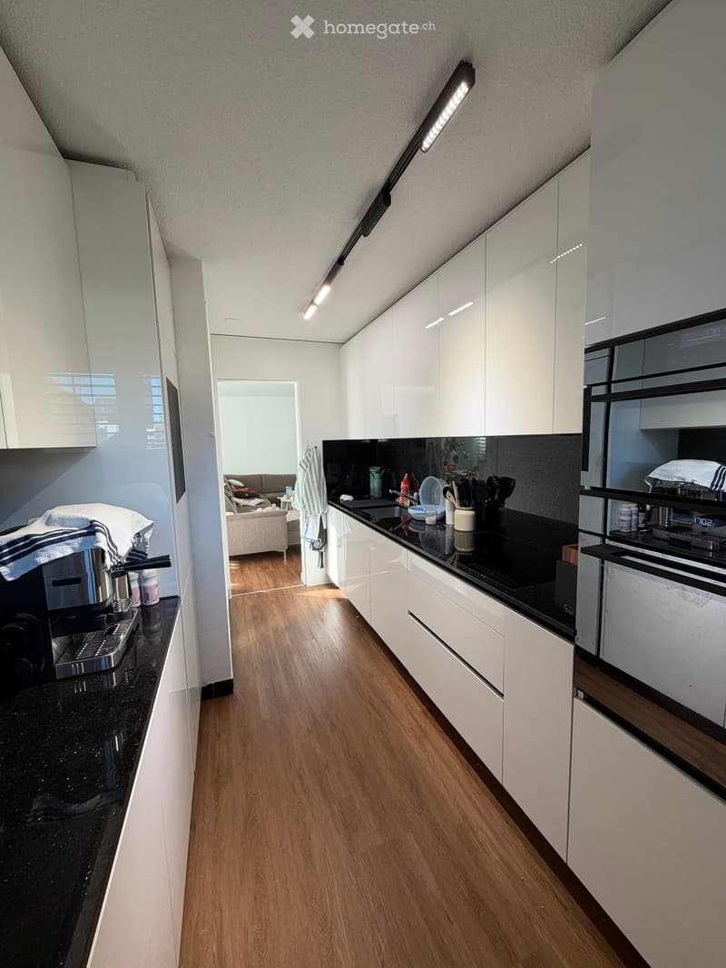 Modern kitchen with white cabinets, black countertops, and wooden flooring. Visible appliances include a coffee maker, oven, and fridge. A small living area with a sofa is visible through an open doorway.