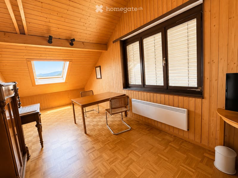 Cozy wooden room with slanted ceiling, large windows with blinds, a small table with chairs, radiator, and a TV screen on a stand.