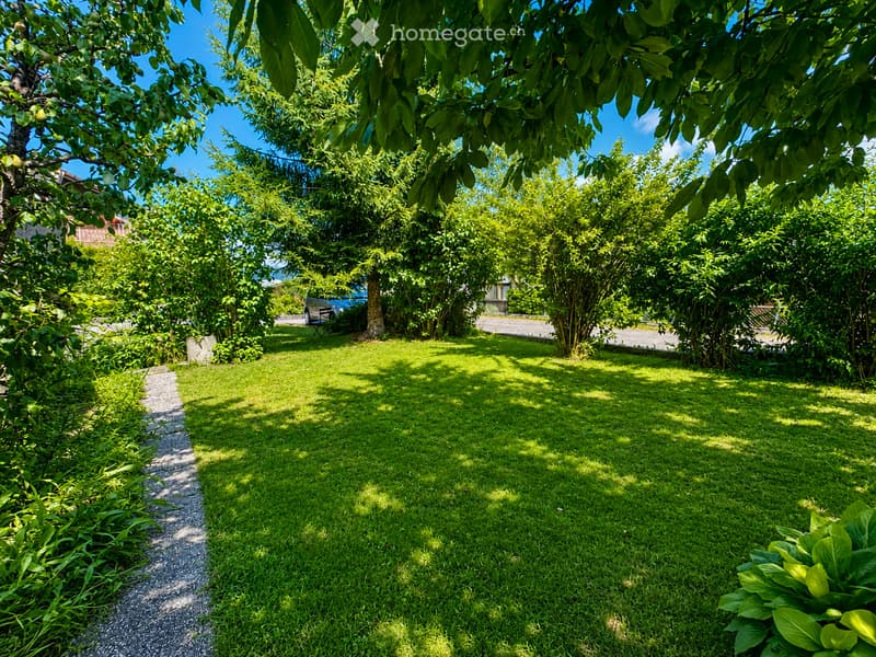 A well-maintained garden with lush green grass, trees, and a gravel pathway. The area is shaded by large trees, creating dappled sunlight on the lawn.
