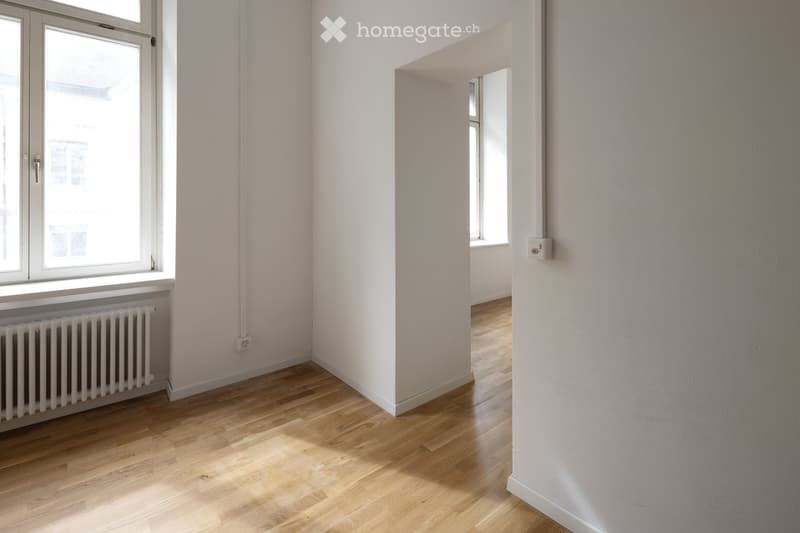 Empty room with light wooden flooring, white walls, and a radiator under a window. No furniture or equipment is present.