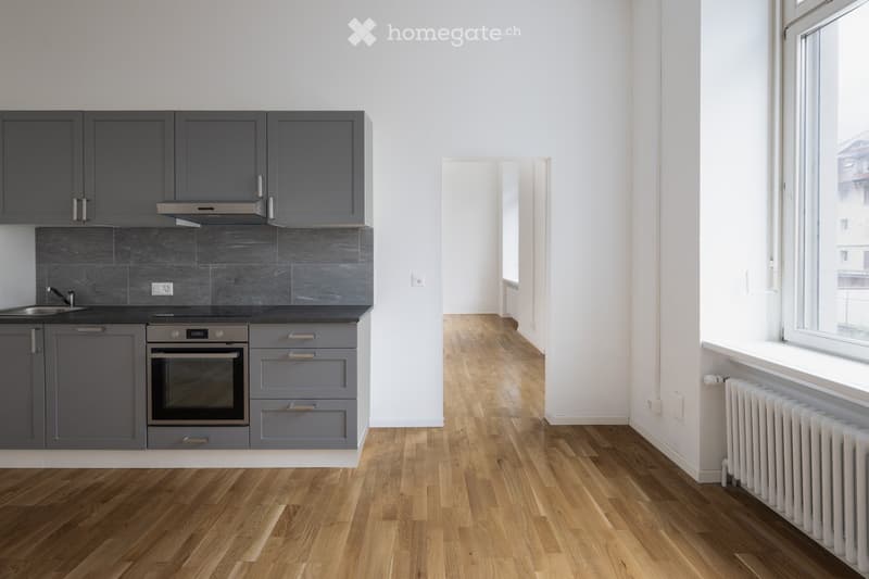 Modern kitchen with grey cabinets, stainless steel oven, sink, and faucet. Light wooden flooring and white walls.