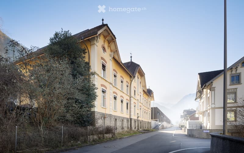 A multi-story building with yellow facade and white trim, featuring arched windows and decorative roofline, situated on a quiet street with a tree and distant mountain view.