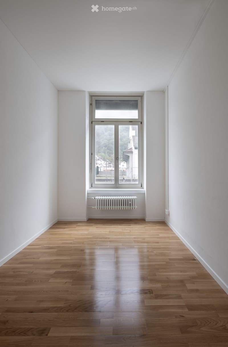 Empty room with light wooden flooring, white walls, and a large window with a radiator below it.