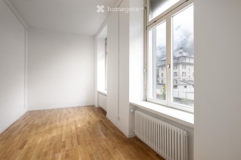 Empty room with light wooden flooring, white walls, and large windows offering a view of buildings and a cloudy sky. Radiators are visible beneath the windows.
