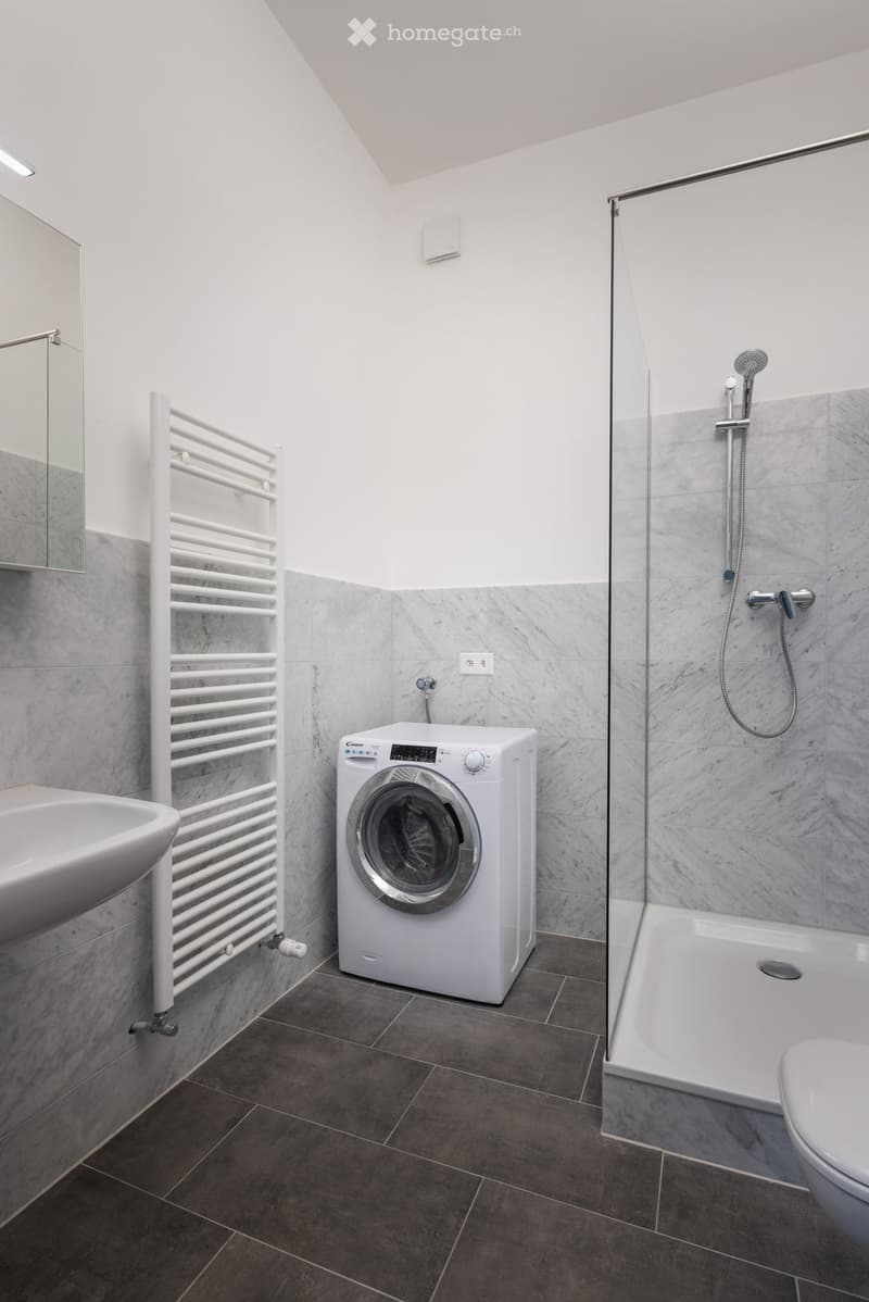 Modern bathroom with a white washing machine, sink, toilet, shower, and a white radiator. The walls are tiled in a light grey marble pattern, and the floor is dark grey tiles.