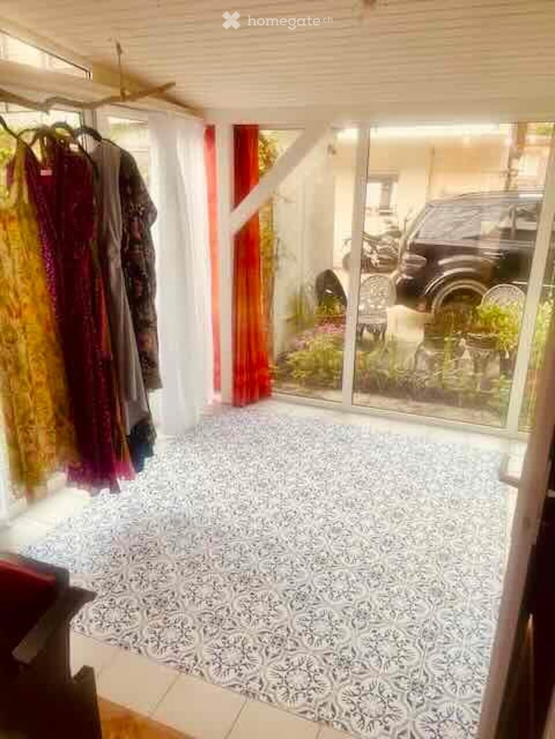 Bright room with patterned floor, large glass doors leading to a garden, clothing rack with garments, and a chair visible through the glass.