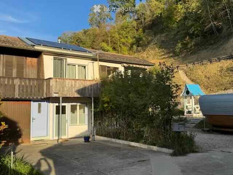 Two-story house with solar panels on the roof, wooden balcony, and surrounding greenery.