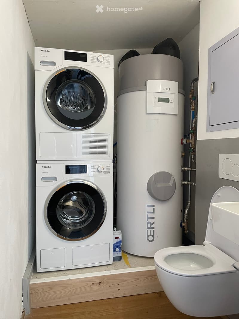 Stacked washing machine and dryer, gas water heater, and white toilet in the corner.