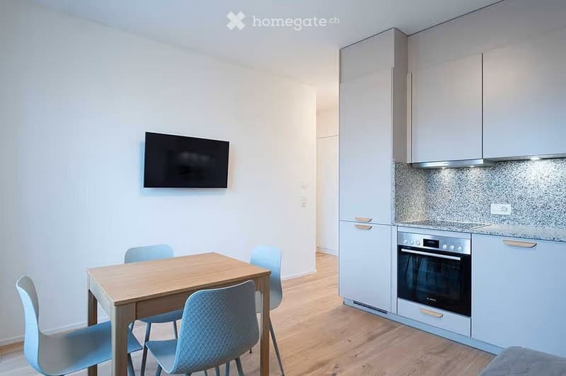 A modern, well-lit room with a wooden dining table and four light blue chairs, a flat-screen TV mounted on the wall, and a fitted kitchen area with light grey cabinets and a built-in oven.