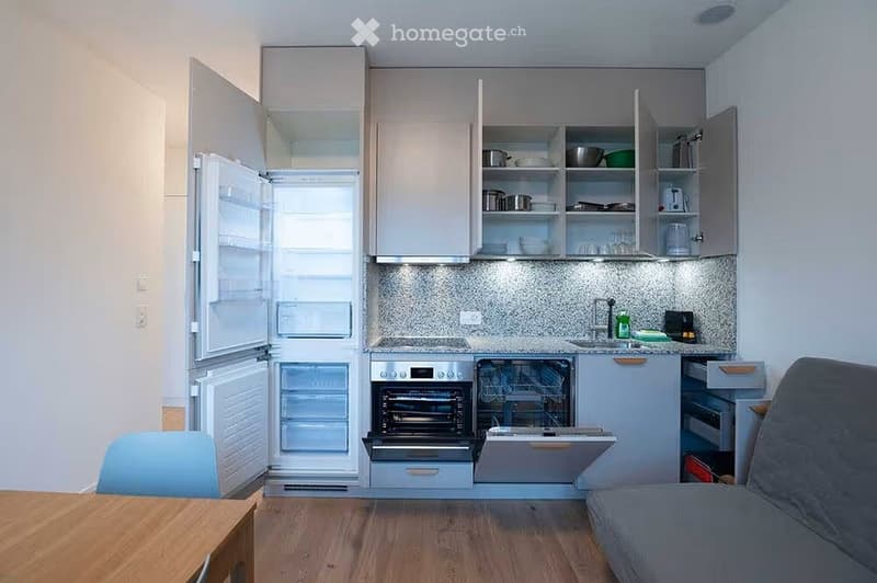 Modern, compact kitchen and living area with grey cabinets, stainless steel appliances, a sofa, a wooden table, and a blue chair.