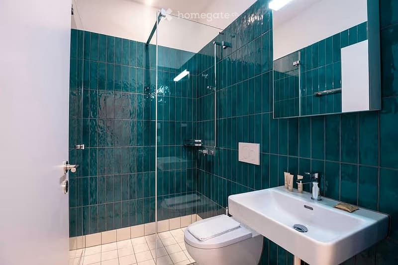 Modern bathroom with green tiled walls, a glass-enclosed shower, a white sink with a faucet, and a toilet. A mirror is mounted above the sink, and a small cabinet is visible on the wall.