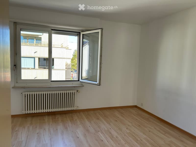 Empty room with light wooden flooring, white walls, and large windows with open shutters, revealing a view of a neighboring building and outdoor area.