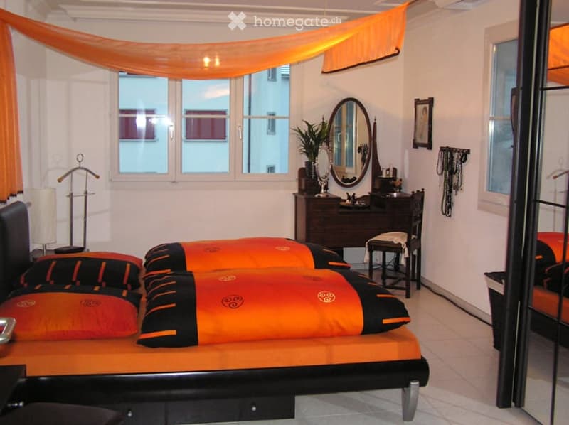 A well-lit bedroom with a large bed featuring orange and black pillows, a wooden desk with a mirror, a chair, a plant, and a clothing rack. The room has large windows with orange drapes and a cozy ambiance.