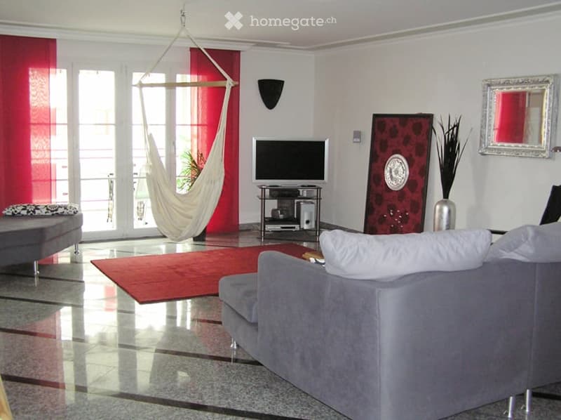 Modern living room with large windows and balcony access, featuring a grey sofa, coffee table, hammock, television, and decorative art pieces.