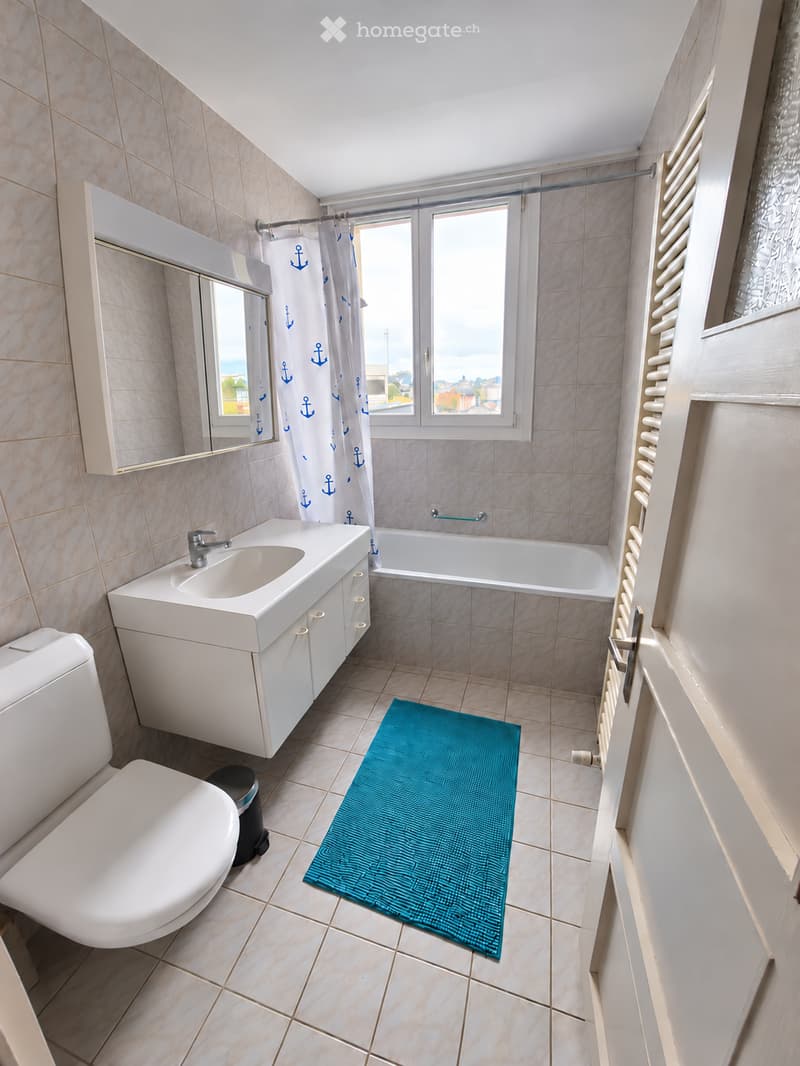Modern bathroom with beige tiles, white sink, toilet, bathtub, mirror, and a blue bath mat.