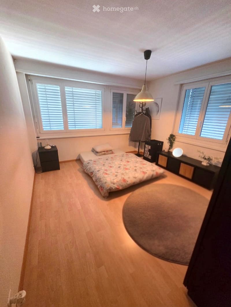 A modern, well-lit bedroom with wooden flooring, a large bed with floral bedding, nightstands, a hanging lamp, and a round rug.