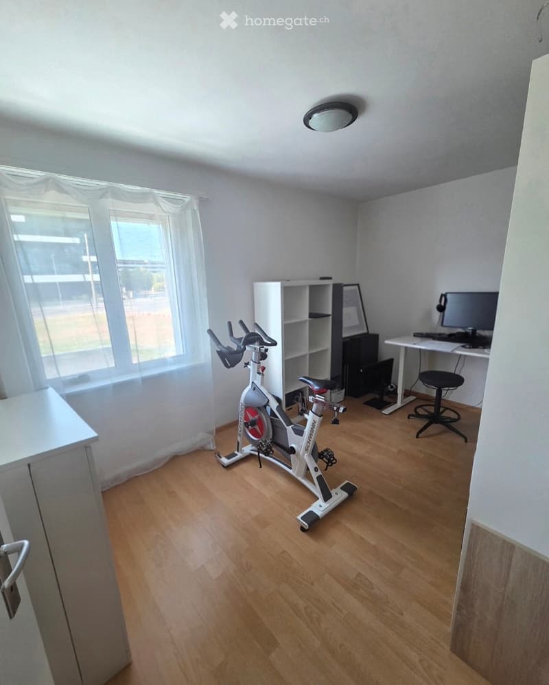 A well-lit room with wooden flooring, featuring an exercise bike, a desk with a computer, a chair, and a shelf.