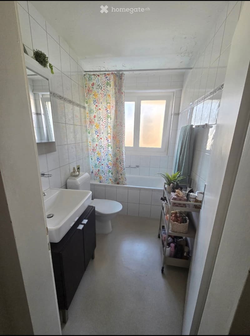 Bathroom with white tiles, a sink, toilet, bathtub with a floral shower curtain, a mirror, and a small shelf with plants and items.